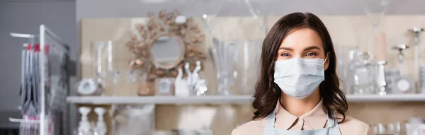 Brünette Blumenhändlerin in medizinischer Maske blickt in die Kamera mit verschwommenem Gestell von Vasen auf dem Hintergrund, Banner — Stockfoto