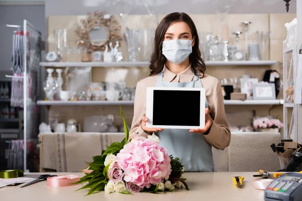 Fleuriste en masque médical montrant tablette numérique près de la table avec bouquet, outils et rubans décoratifs sur fond flou — Photo de stock