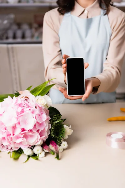 Vista ritagliata del fiorista che mostra smartphone con schermo bianco vicino al bouquet sulla scrivania su sfondo sfocato — Foto stock