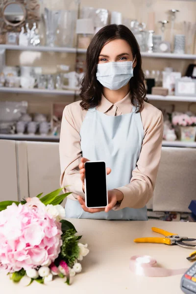 Floristería en máscara médica que muestra el teléfono inteligente con pantalla en blanco cerca del ramo en el escritorio con bastidores borrosos de jarrones en el fondo - foto de stock