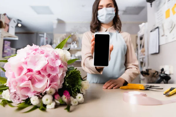 Vista de perto do smartphone nas mãos do florista perto do buquê na mesa com ferramentas na loja de flores no fundo borrado — Fotografia de Stock