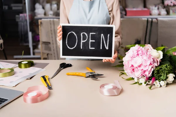 Cropped view of florist holding chalkboard with open lettering near fresh bouquet on desk on blurred background — Stock Photo