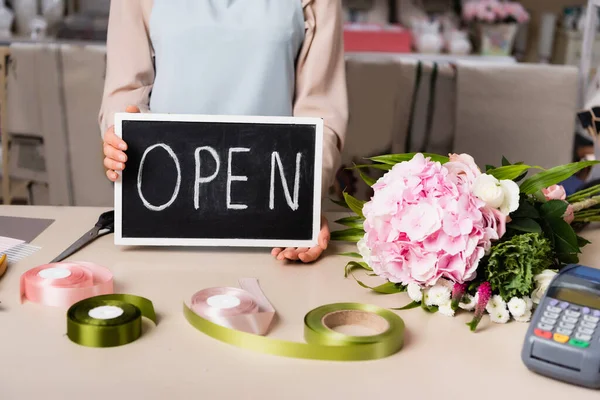 Cropped view of florist holding chalkboard with open lettering near bouquet on desk on blurred background — Stock Photo