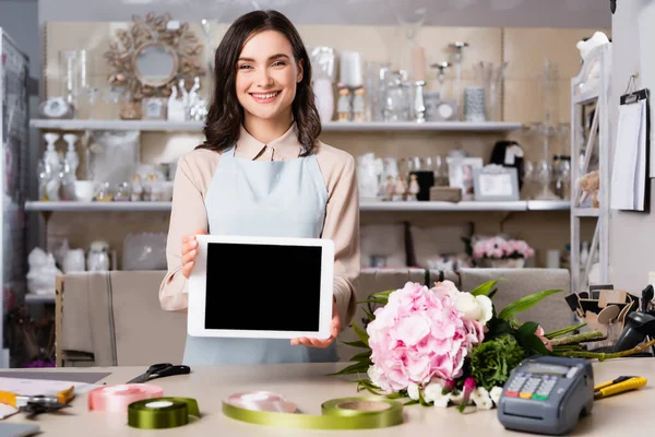 Happy florist showing digital tablet near bouquet on desk with blurred racks of vases on background — Stock Photo