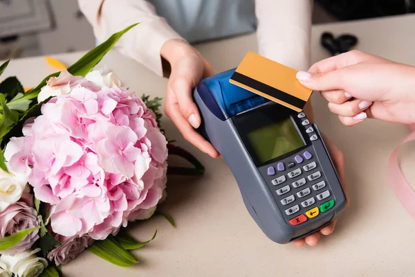 Vista cortada do cliente que paga com cartão de crédito pelo terminal nas mãos do florista perto de hydrangea florescendo na mesa — Fotografia de Stock