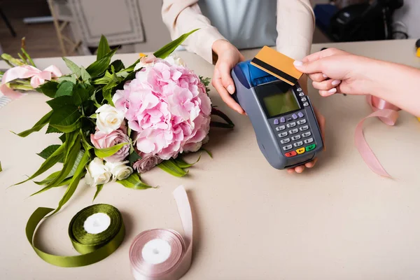 Vista recortada del cliente pagando con tarjeta de crédito por terminal en manos de florista cerca de ramo en escritorio con cintas decorativas - foto de stock