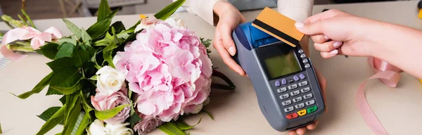 Vue recadrée du client payant avec carte de crédit par terminal dans les mains du fleuriste près du bouquet frais sur le bureau, bannière — Photo de stock