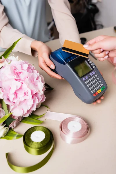 Ausgeschnittene Ansicht des Kunden, der mit Kreditkarte per Terminal in den Händen eines Blumenhändlers in der Nähe von Hortensien und dekorativen Bändern auf dem Schreibtisch bezahlt — Stockfoto