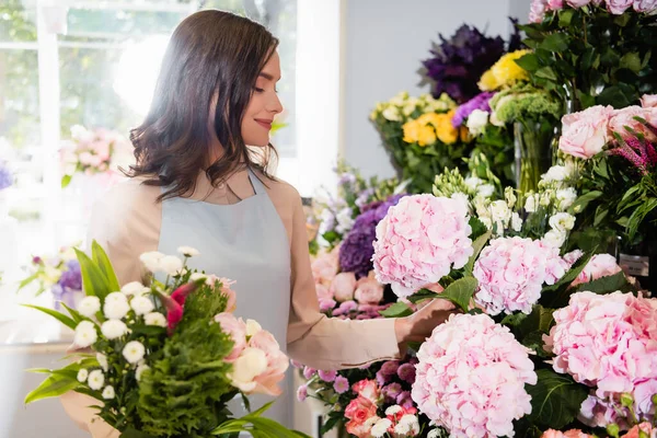Smiling florist with bouquet taking hydrangea in flower shop with blurred window on background — Stock Photo