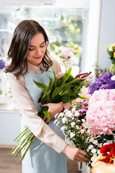 Florista sonriente tomando rama de crisantemo, mientras que la recolección de ramo cerca de la gama de flores con fondo borroso - foto de stock