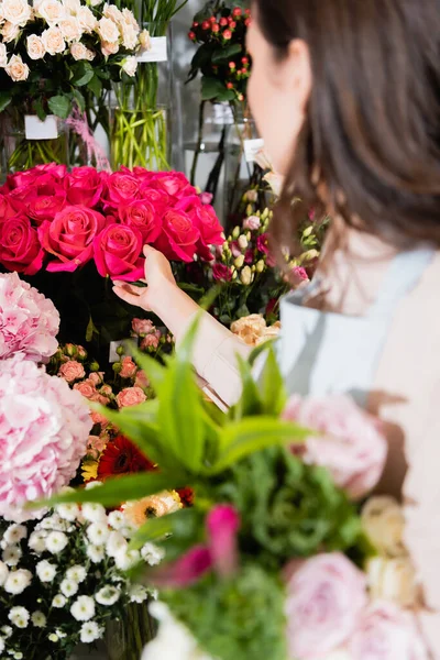 Brunette florist touching rouses in vase on rack of flowers with blurred bouquet on foreground — Stock Photo