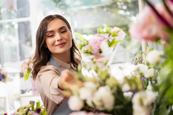 Florista feminino feliz com a mão estendida olhando para flores borradas em primeiro plano — Fotografia de Stock