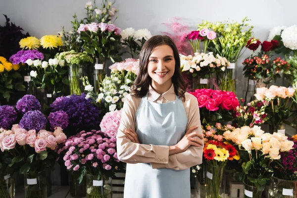Усміхнений жіночий флорист з схрещеними руками дивиться на камеру, стоячи біля стійок з квітами на фоні — стокове фото