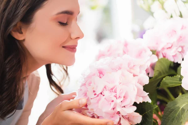 Side view of smiling woman with closed eyes smelling hydrangea on blurred background — Stock Photo