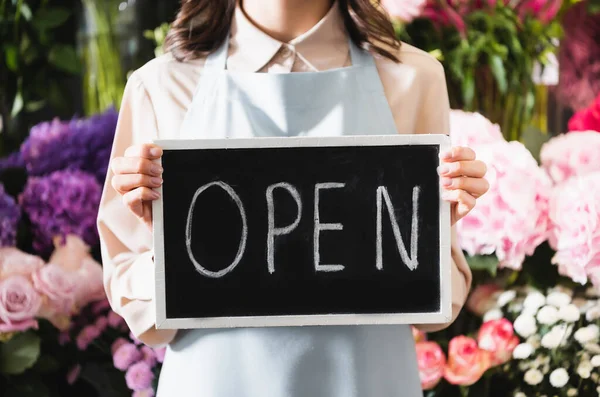 Cropped view of florist holding chalkboard with open lettering with blurred range of flowers on background — Stock Photo