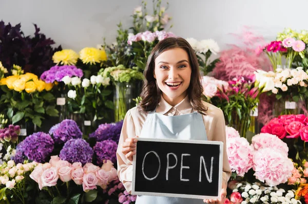 Emocionada florista femenina mirando a la cámara mientras sostiene la pizarra con letras abiertas con bastidores de flores en el fondo - foto de stock