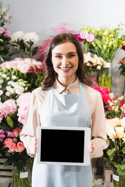Vista frontal de la floristería feliz mirando a la cámara mientras sostiene la tableta digital con bastidores borrosos de flores en el fondo - foto de stock