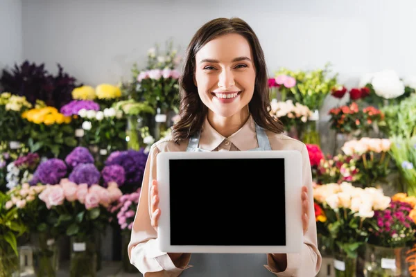 Fröhliche Floristin zeigt digitales Tablet mit verschwommenem Blumengestell auf Hintergrund — Stockfoto