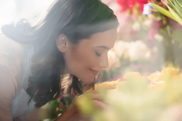 Vue latérale du fleuriste féminin souriant avec les yeux fermés sentant la fleur sur le premier plan flou — Photo de stock