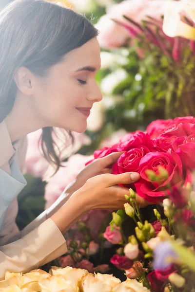 Vista lateral de la floristería femenina feliz con los ojos cerrados oliendo rosas con la gama borrosa de flores en el fondo - foto de stock
