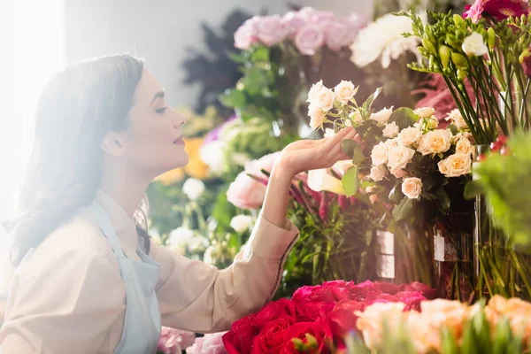 Side view of happy female florist caring about roses near flower range on blurred foreground — Stock Photo