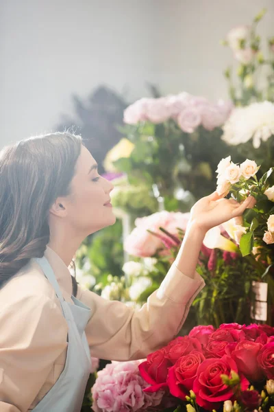 Seitenansicht eines lächelnden brünetten Blumenhändlers, der sich um Rosen auf Blumenständern auf verschwommenem Hintergrund kümmert — Stockfoto