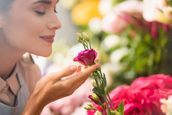 Happy female florist with closed eyes smelling eustoma flower with blurred flowers on background — Stock Photo