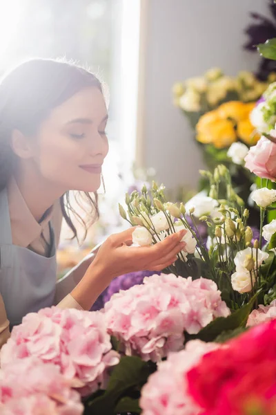 Happy florist with closed eyes smelling eustoma flowers near hydrangeas with blurred window on background — Stock Photo