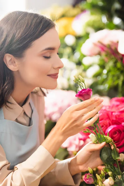 Brünette Floristin mit geschlossenen Augen riecht Eustoma-Blume in der Nähe von Rosen auf verschwommenem Hintergrund — Stockfoto