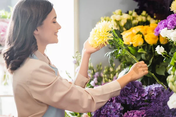 Vue latérale du fleuriste heureux se souciant de jaune aster près de la gamme de fleurs sur fond flou — Photo de stock