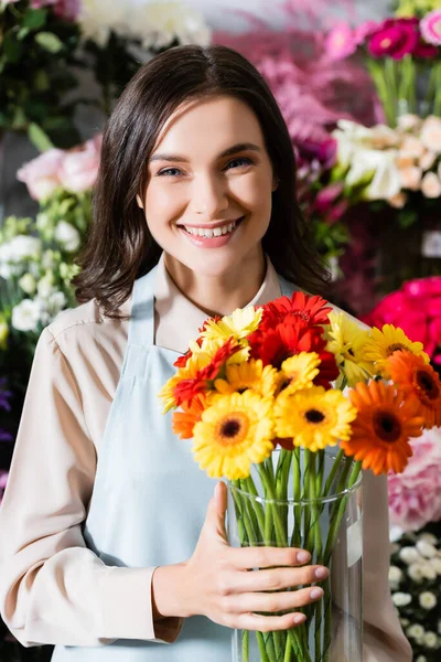 Sorrindo florista fêmea olhando para a câmera enquanto segurando vaso com gerberas com gama turva de flores no fundo — Fotografia de Stock