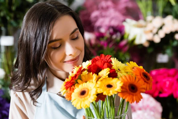 Brunette florist with closed eyes smelling gerberas in vase near blurred range of flowers on background — Stock Photo