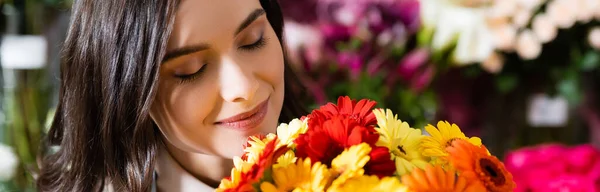 Florista sorridente com olhos fechados cheirando gerberas com gama turva de flores no fundo, banner — Fotografia de Stock