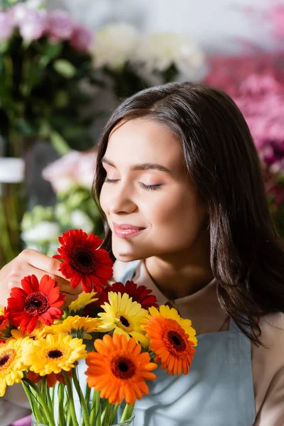 Lächelnde Floristin mit geschlossenen Augen, die Gerberas mit verschwommenen Blumen auf dem Hintergrund riecht — Stockfoto