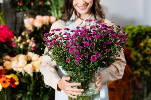 Vista recortada de floristería femenina sosteniendo jarrón con crisantemos púrpura cerca de borrosa gama de flores en el fondo - foto de stock