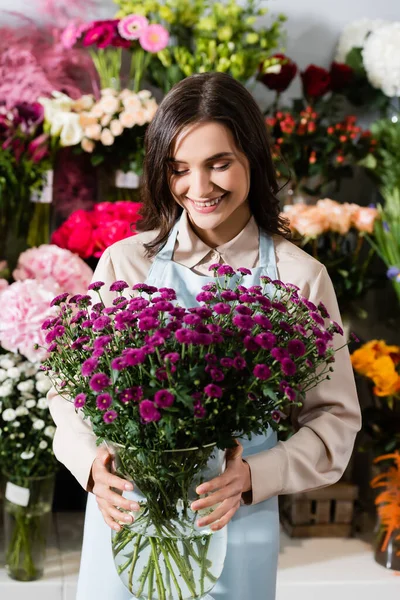 Vista frontal da florista feminina feliz olhando para crisântemos roxos em vaso perto de prateleiras borradas de flores no fundo — Fotografia de Stock