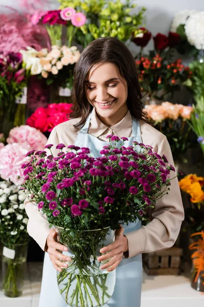 Vista frontal da florista feminina feliz olhando para crisântemos roxos em vaso perto de prateleiras borradas de flores no fundo — Fotografia de Stock