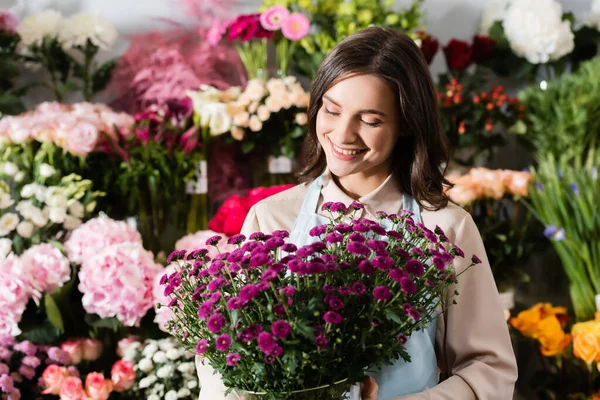 Вид спереди улыбающейся флористки, смотрящей на фиолетовые хризантемы в вазе возле размытых вешалок с цветами на заднем плане — стоковое фото