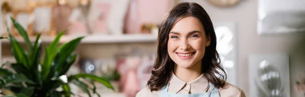 Retrato de florista feminino sorridente olhando para a câmera com loja de flores borradas no fundo, banner — Fotografia de Stock