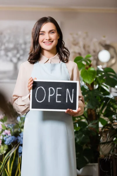 Front view of smiling female florist holding chalkboard with open lettering on blurred foreground — Stock Photo