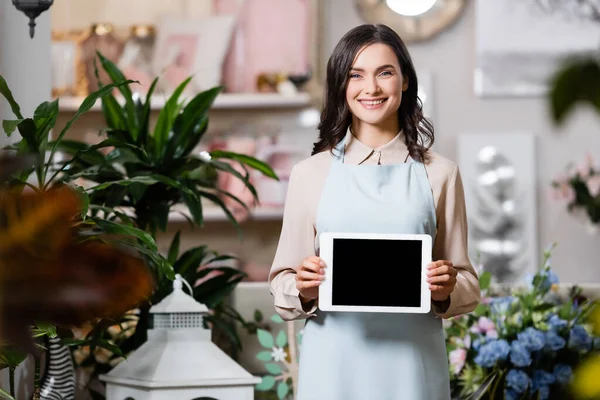 Glückliche Floristin blickt in die Kamera, während sie im Blumenladen ein digitales Tablet im verschwommenen Vordergrund hält — Stockfoto