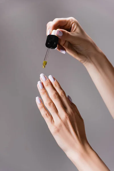 Vista recortada de la mujer aplicando aceite de pipeta en las uñas aisladas en gris - foto de stock