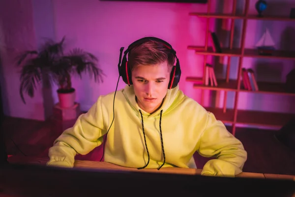 Young gamer in headset using computer on blurred foreground at home — Stock Photo