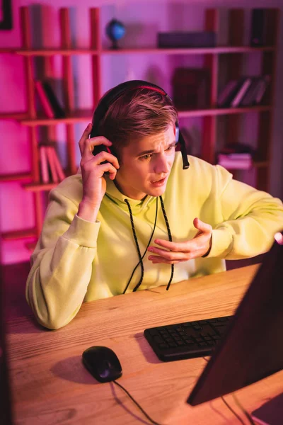 Jugador usando auriculares mientras habla durante el videojuego cerca de la computadora en primer plano borroso - foto de stock