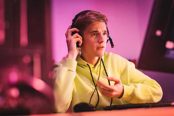 Jeune joueur avec la main sur le casque regardant l'écran d'ordinateur sur le premier plan flou à la maison — Photo de stock