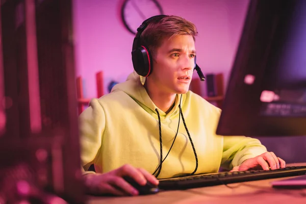 Concentrated gamer in headset using computer keyboard and mouse on blurred foreground — Stock Photo