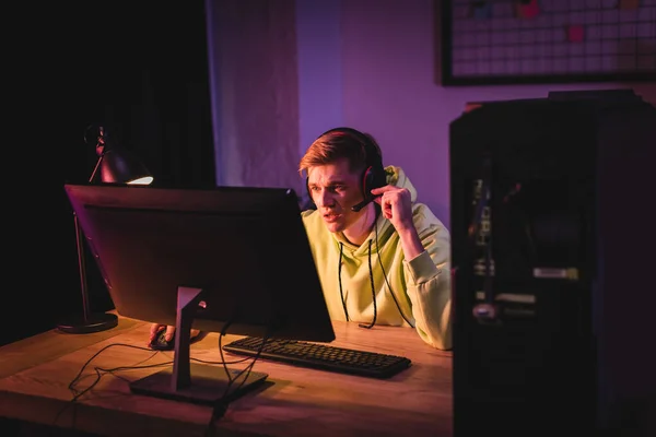 Gamer concentrado en auriculares jugando en la computadora cerca de la lámpara en la mesa - foto de stock