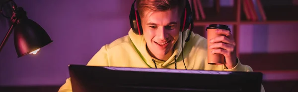 Jogador alegre em fone de ouvido segurando café para ir e olhando para monitor de computador em primeiro plano desfocado, banner — Fotografia de Stock