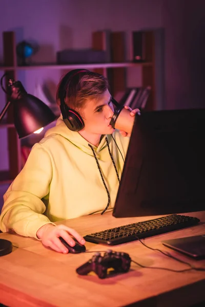 Gamer en auriculares bebiendo café para ir mientras usa la computadora cerca de joystick en primer plano borroso - foto de stock