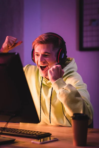 Hombre alegre en auriculares mostrando sí gesto mientras juega videojuego en el ordenador cerca de café para llevar en primer plano borrosa - foto de stock